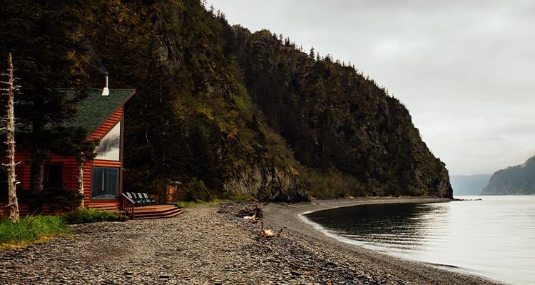 A wooden lodge below cliffs on a rocky beach by the sea.