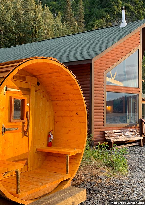 A wooden sauna next to a wooden lodge below forested cliffsides.