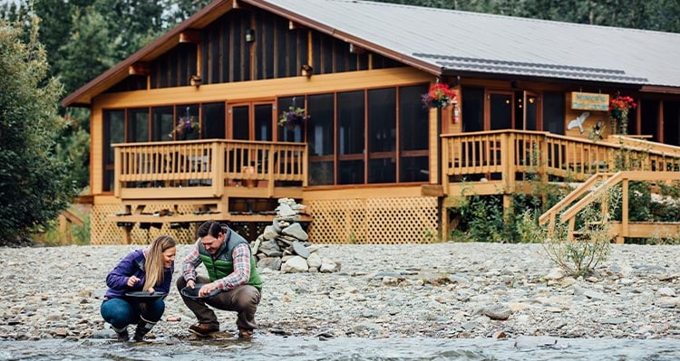 A couple look at the rocks in the river outside of a wooden cabin.