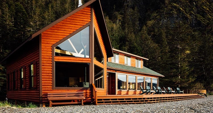 A log cabin hotel sits on a stone beach, forest behind.