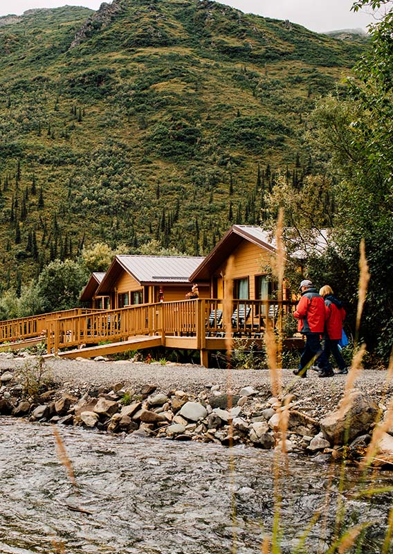 A log cabin hotel in lush green landscape, sits at a rivers edge, a couple walks along the river.