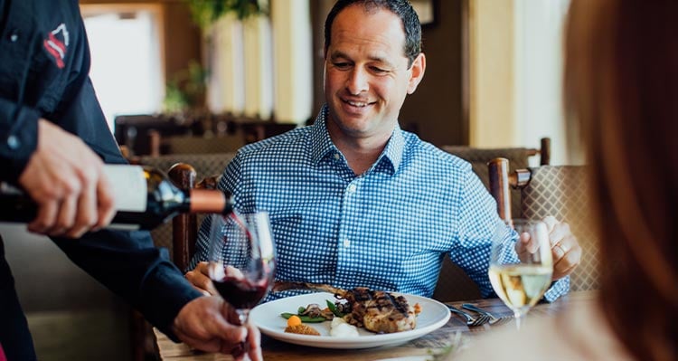 A man eats dinner at a table with the server presenting a bottle of wine.