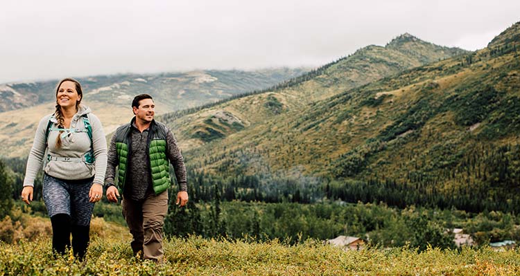 A man and a woman hike in the daytime through tundra grass.