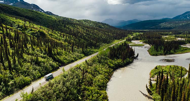 A bus drives alongside a river with a winding shoreline and forest to the other side of the road.