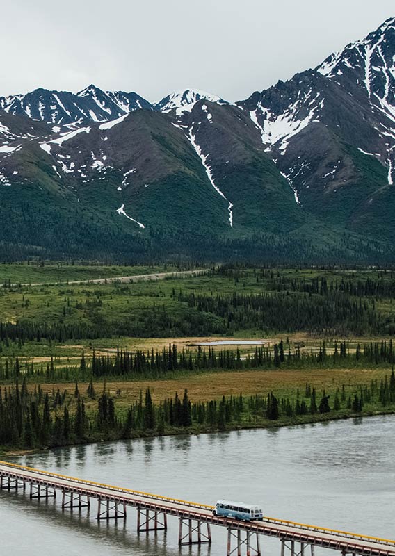 A blue bus drives across a long bridge over water with mountains and trees in the background.