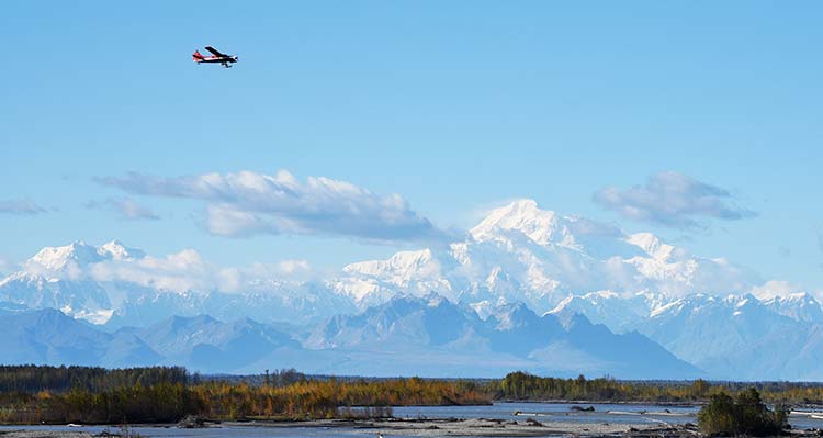 A small plane flies over a river bed during the day with mountains in the background