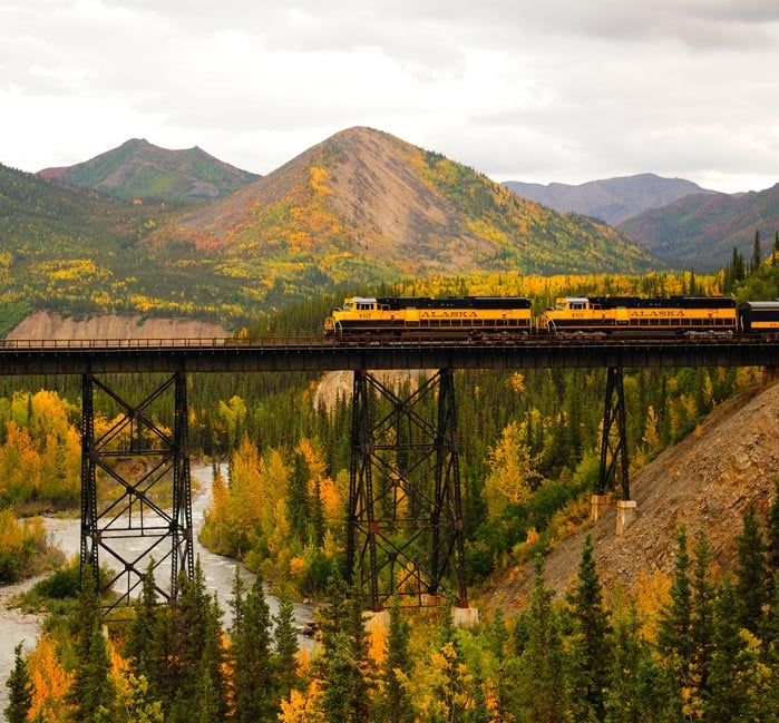 A blue and yellow train moves along a tall bridge over a winding river.