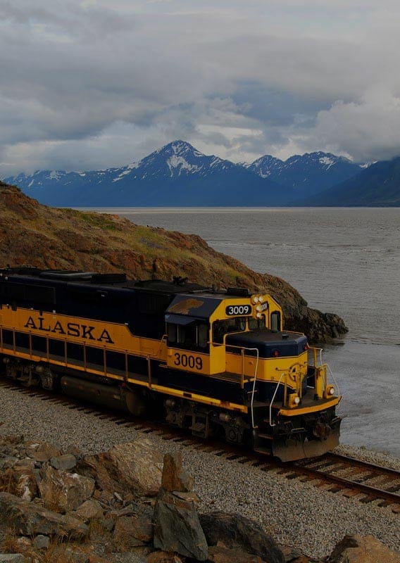 A train travels along a rocky shoreline with mountains in the distance