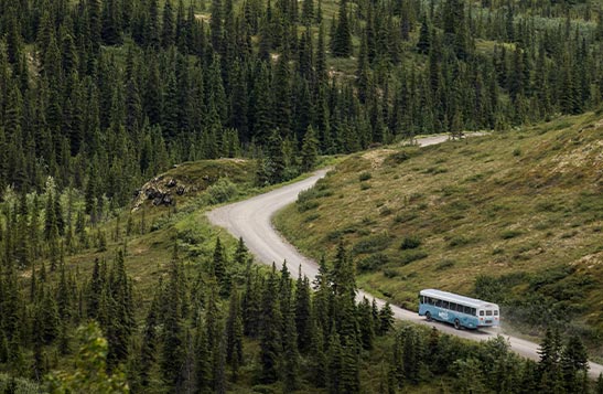 A bus drives down a remote road in sparsely treed terrain and snow covered mountains in the distance