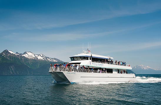 A Kenai Fjords Tour boat full of people cruise thought water with a mountain in the background.