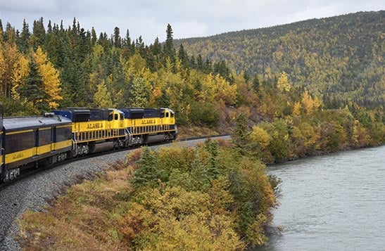 A train rounds a curve through a forest below snow-covered mountains.