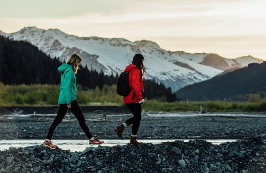 Two people walk on a rocky rivershore below snow-covered mountains.