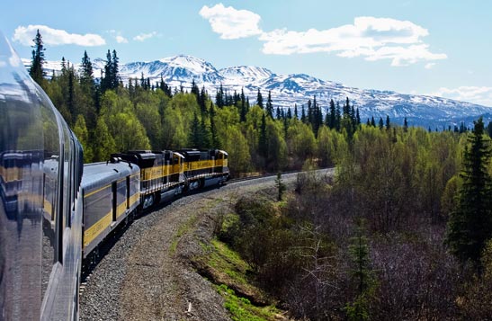 A train rounds a curve through a forest below snow-covered mountains.