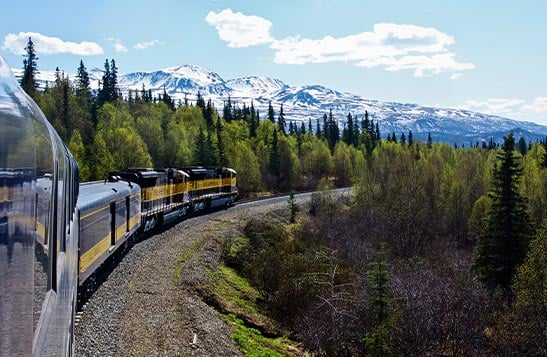 A yellow and blue train moves through forested mountainous terrain