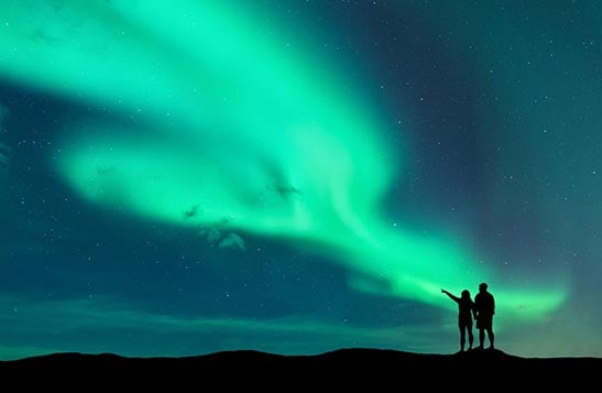 Two people's silhouette's are in front of a large green aurora borealis in the night sky.