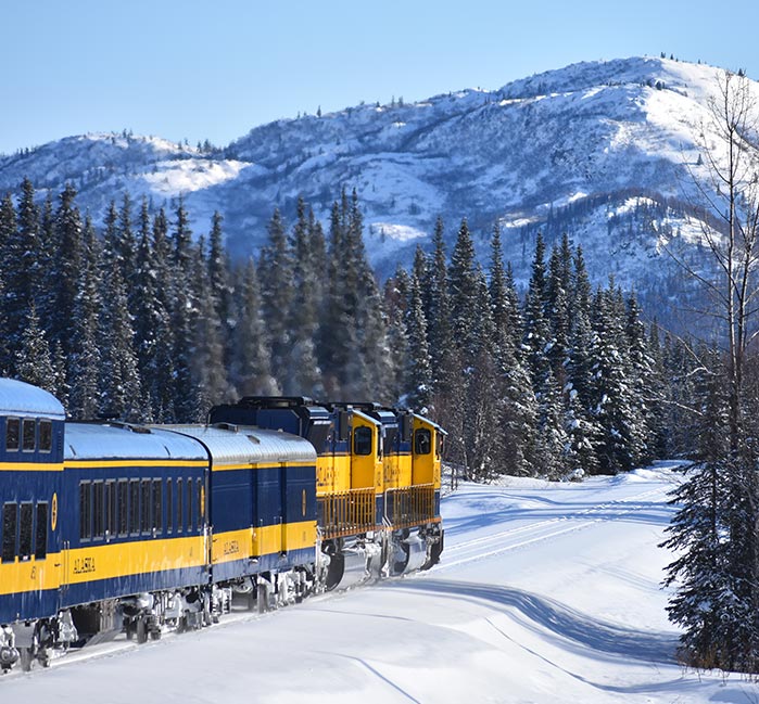 A yellow and blue train goes through the snow with e mountain in the background.