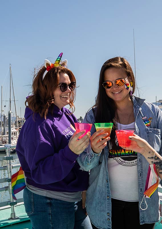 Women stand together wearing rainbow items and cheering drinks.