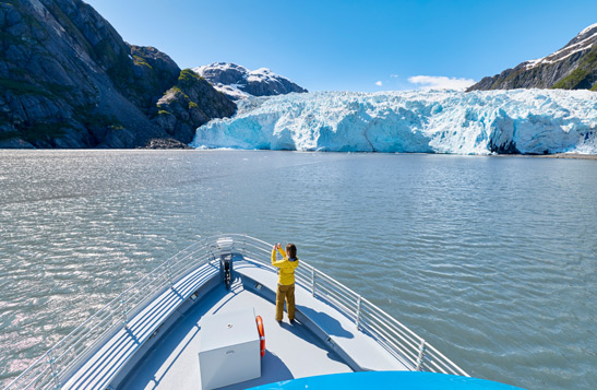 A person standing on the bow of a Kenai Fjords Tour boat taking a photo of the glacier