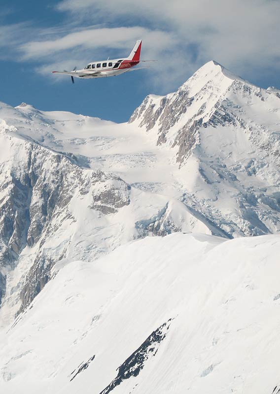 A red and white airplane soars above a snow covered Denali Mountain.