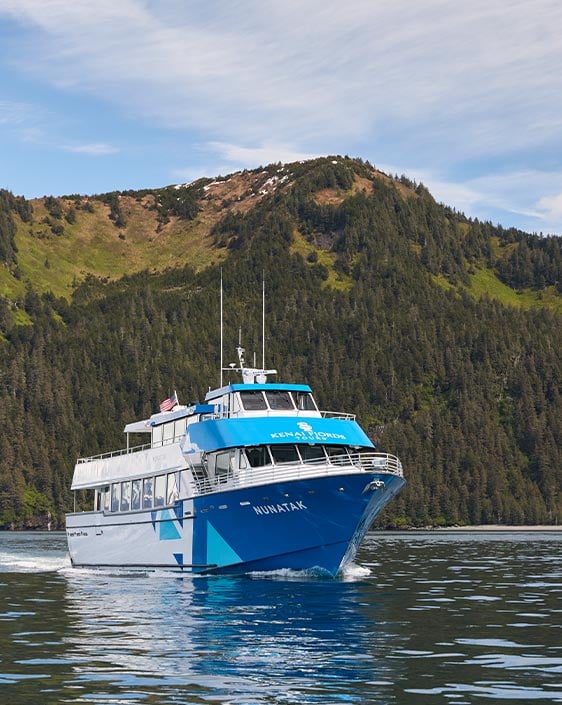 A blue boat cruising on the water near a forested mountainside.