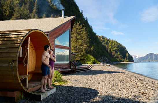 A couple standing outside of a barrel sauna and lodge located on a beach with a mountain view