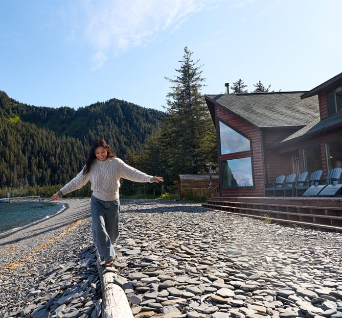 A person walking along a log on the beach in front of Kenai Fjords Wilderness Lodge