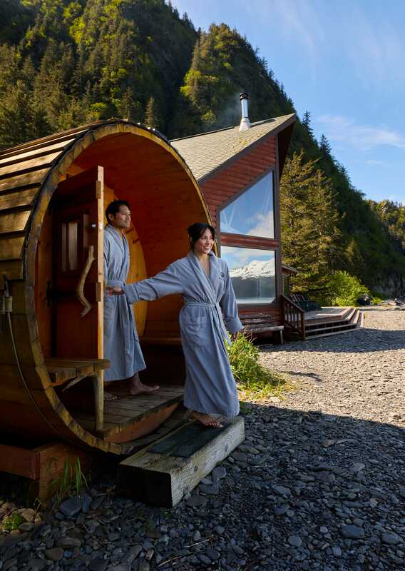 Two people exit a sauna onto a rocky beach by the ocean.