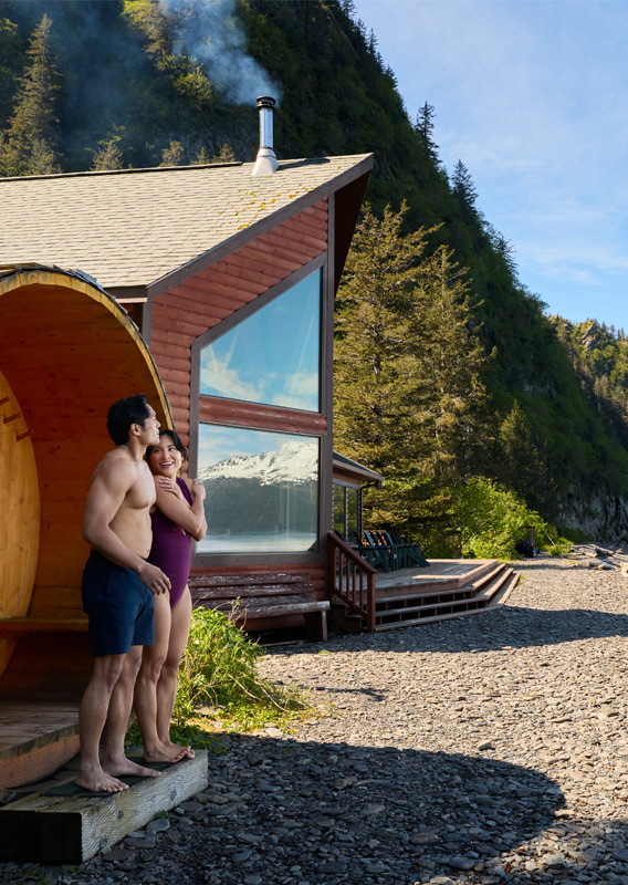 Two people standing outside a sauna beside a lodge on a remote beach.