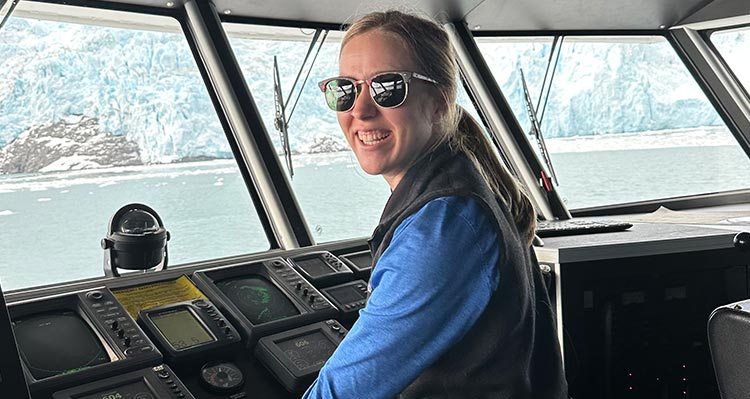 A woman stands at boat controls in a bridge.