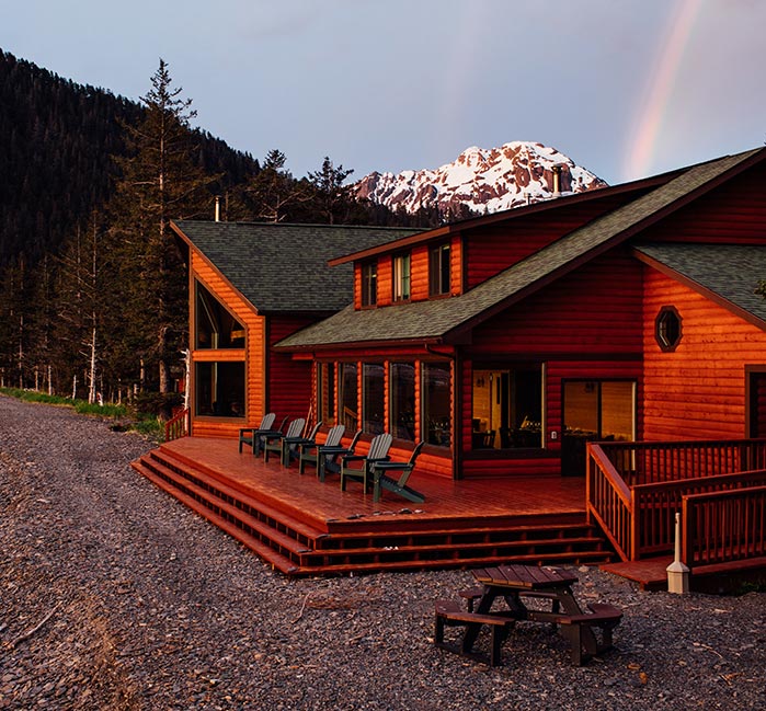 A wooden lodge at a rocky beach below a forested mountainside.