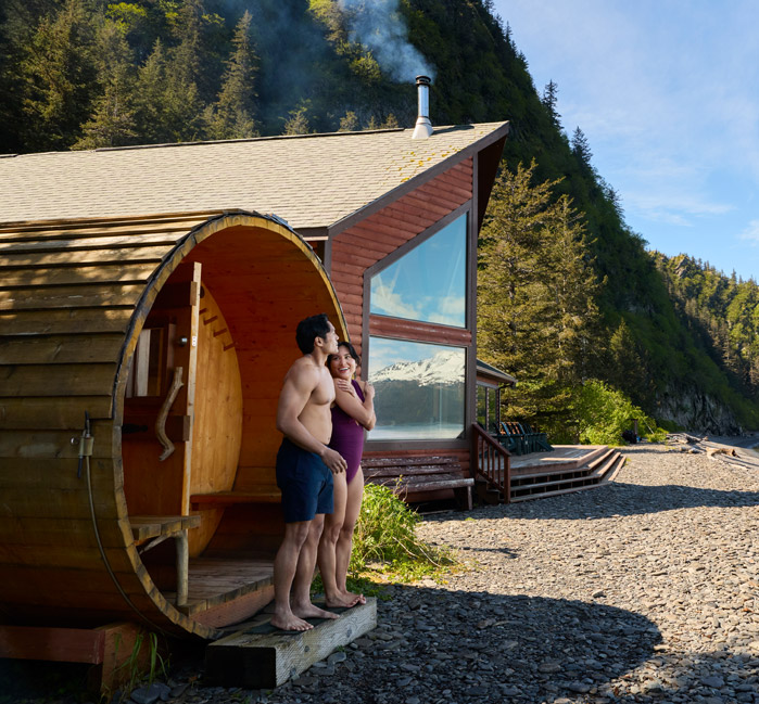 Two people standing outside a barrel sauna beside a lodge on a beach.