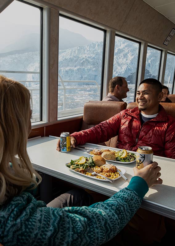 Two people sit at a windowside table for dinner, looking out to a tidewater glacier.