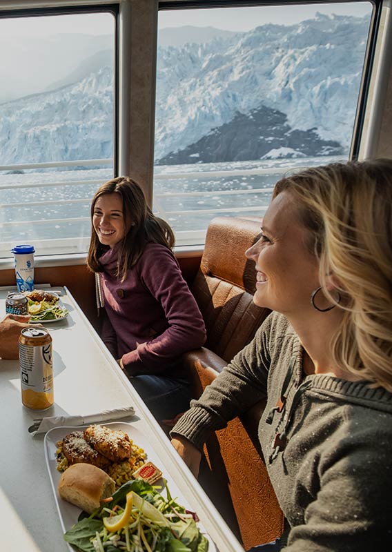 A group of people sit together for dinner at a windowside table looking out to a tidewater glacier.